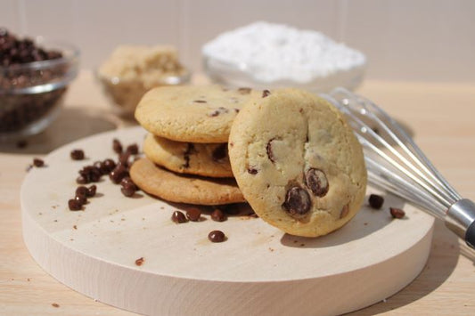 A stack of chocolate chip cookies resting next to a whisk. In the background are small bowls of ingredients used to make cookies.