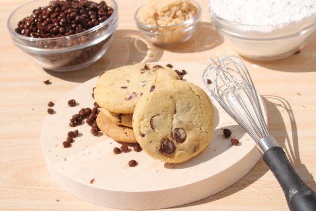 A stack of chocolate chip cookies resting next to a whisk. In the background are small bowls of ingredients used to make cookies.