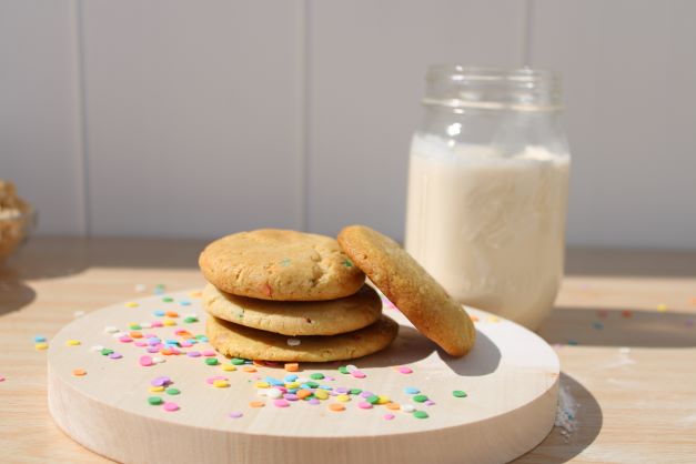 A stack of rainbow sugar cookies resting next to a whisk. In the background are small bowls of ingredients used to make cookies.