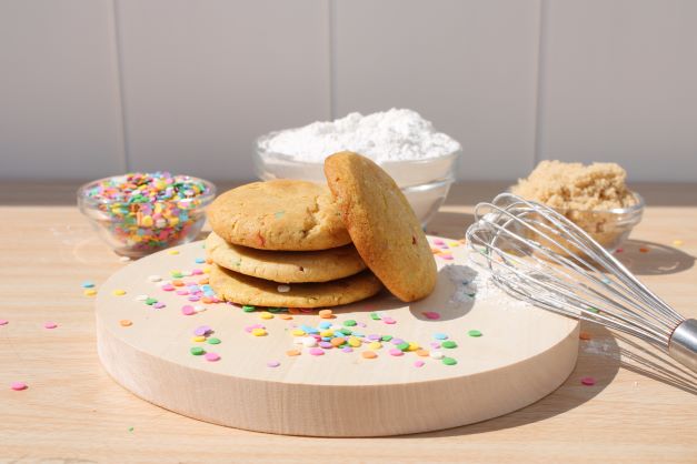 A stack of rainbow sugar cookies resting next to a whisk. In the background are small bowls of ingredients used to make cookies.